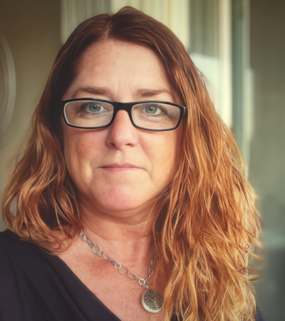 An white woman with reddish-blond hair wearing black eyeglasses looks out confidently.